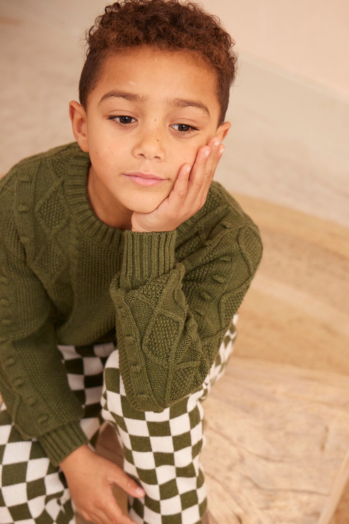 Boy standing against a plaster wall in the Cable Popcorn Jumper in avocado green