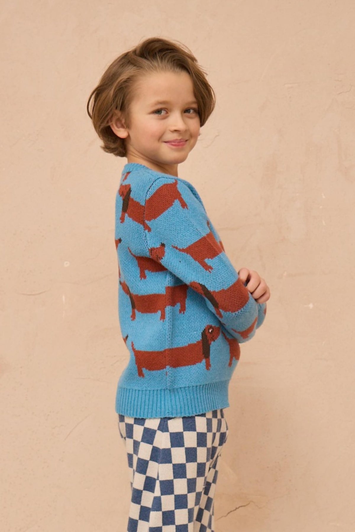 Boy standing against a plaster wall in the Dachshund Jumper in Iceland blue