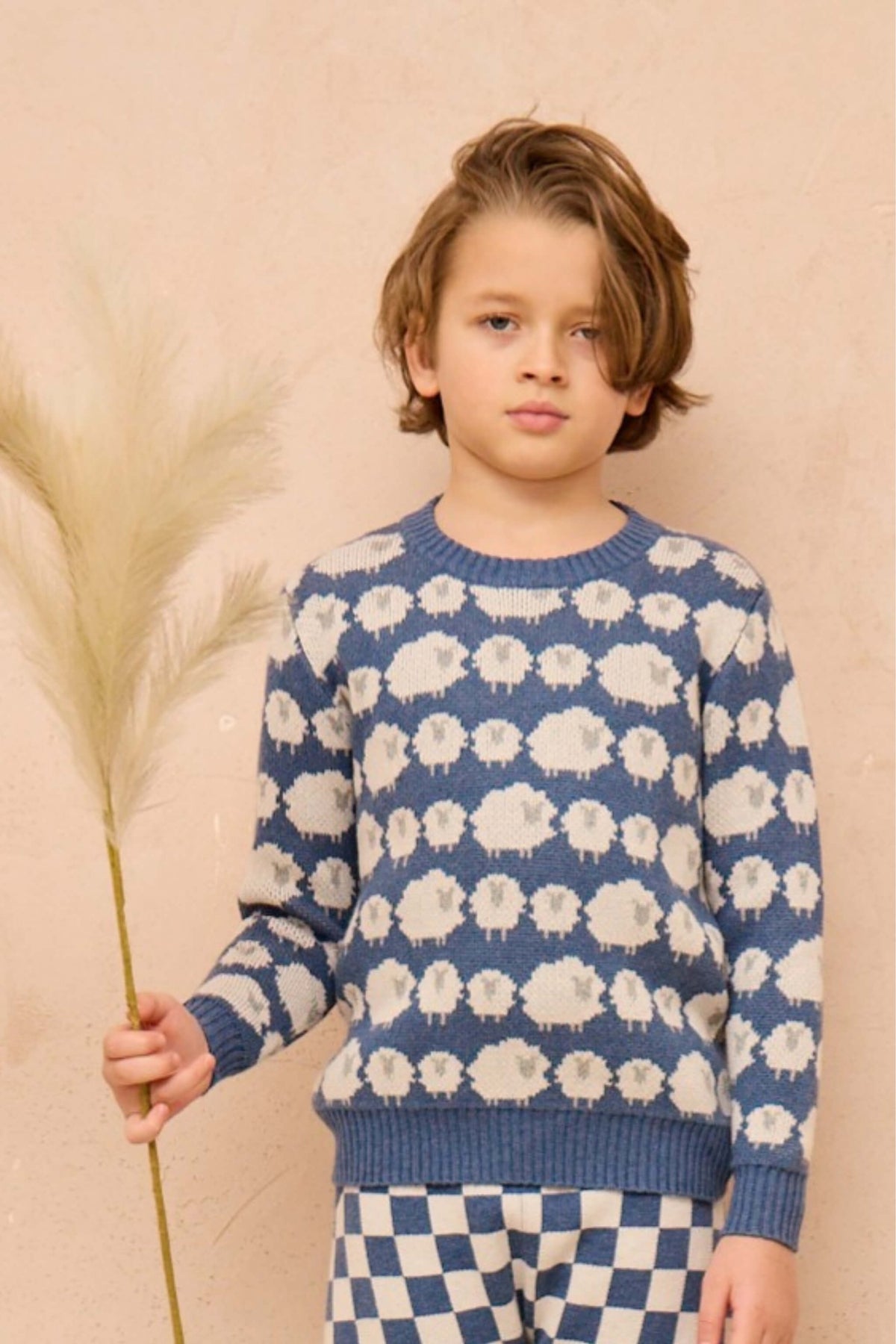 Boy standing against a plaster wall in the Flock of Sheep Jumper in bluestone blue