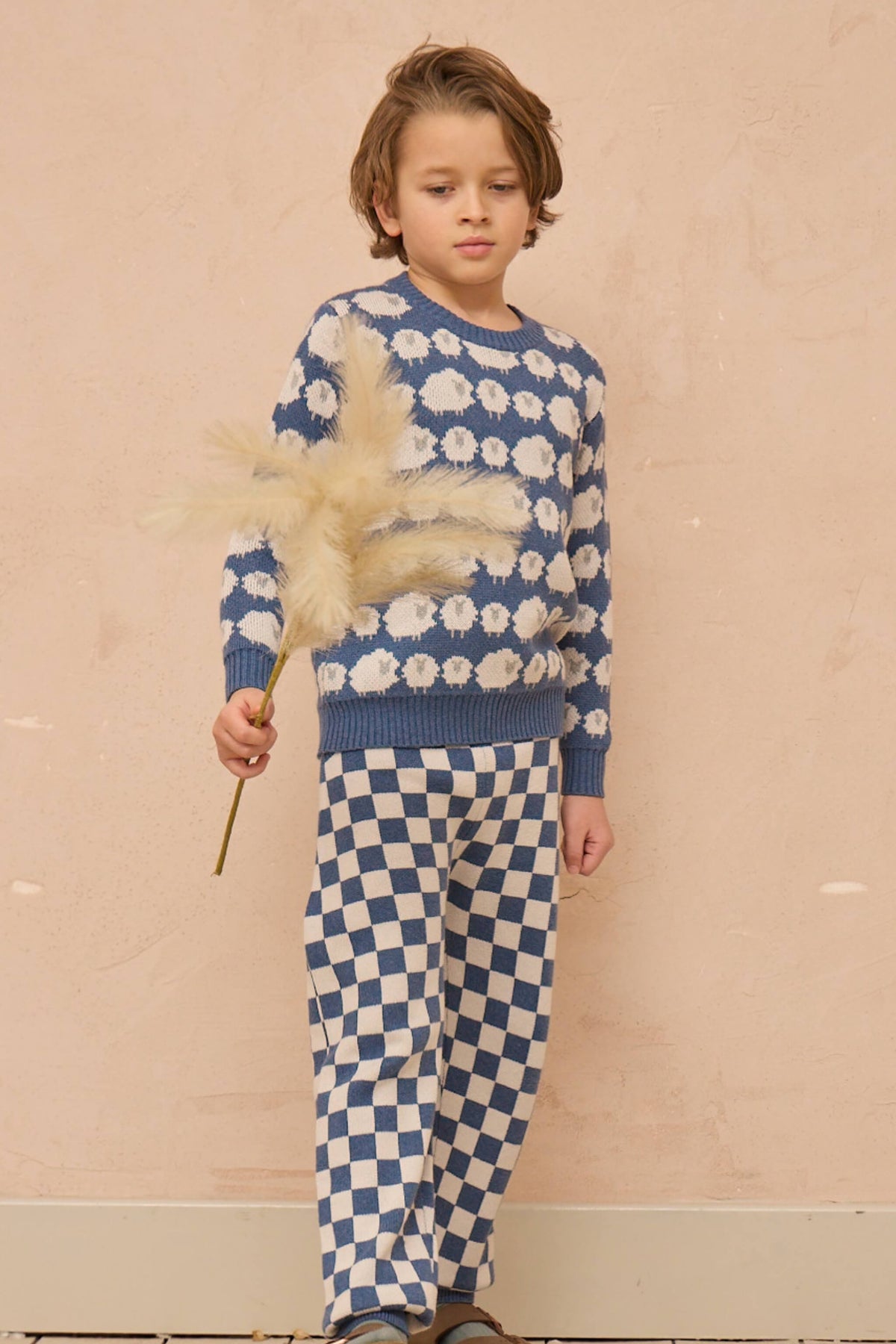 Boy wearing the Flock of Sheep Jumper in bluestone blue with the sheep pattern across the sleeves