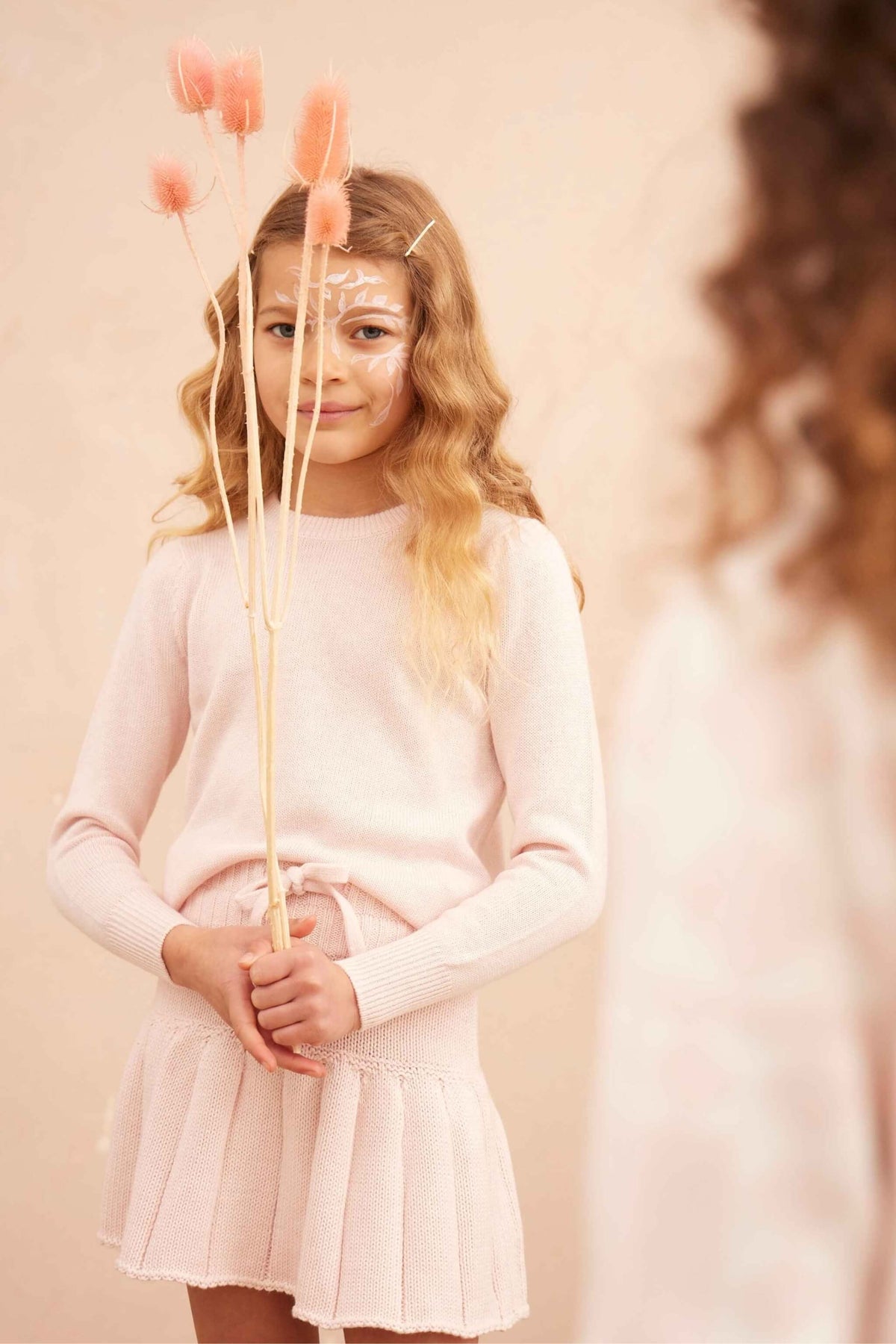 Girl standing against a plaster wall in the Angel Wings Jumper in blush pink