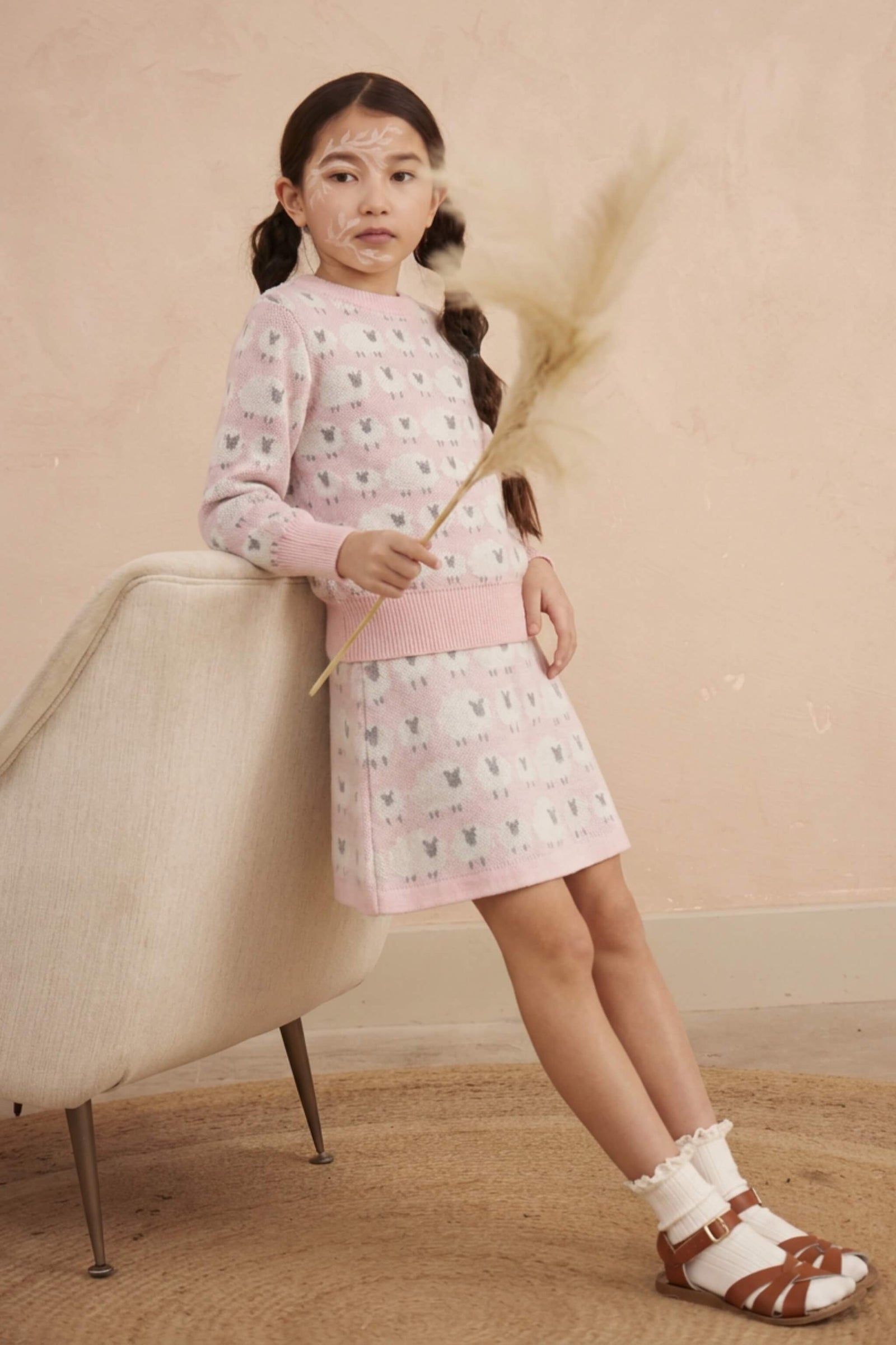 Girl leaning against a chair in the Little Bo Peep jumper and skirt set in rose pink