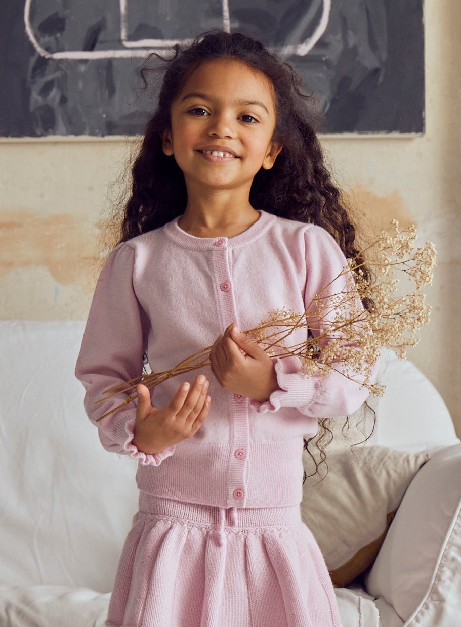This stylish 2 ply cardigan comes with gentle puff sleeves, a long bottom rib to accentuate the waist, and sweet ruffles at the end of the cuff. A fun light weight addition to any winter wardrobe, it is made with the softest cashmere/merino blend from Inner Mongolia, and will keep your Little One stylish and warm all winter long.   Team it up with our matching pleated Tennis skirt in Blossom Pink or a head-to-toe cosy feel. 